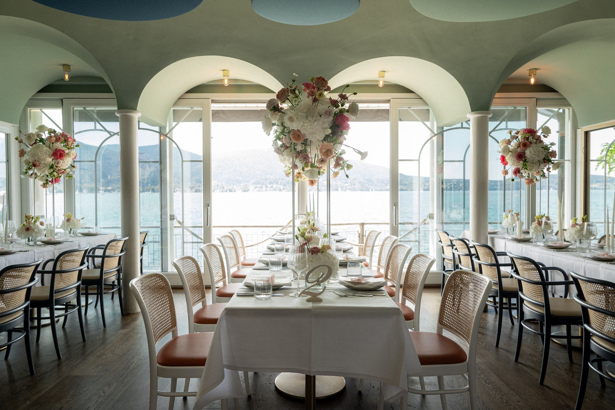 Dinner tables with tall floral centerpieces face Lake Tegernsee through arched windows.