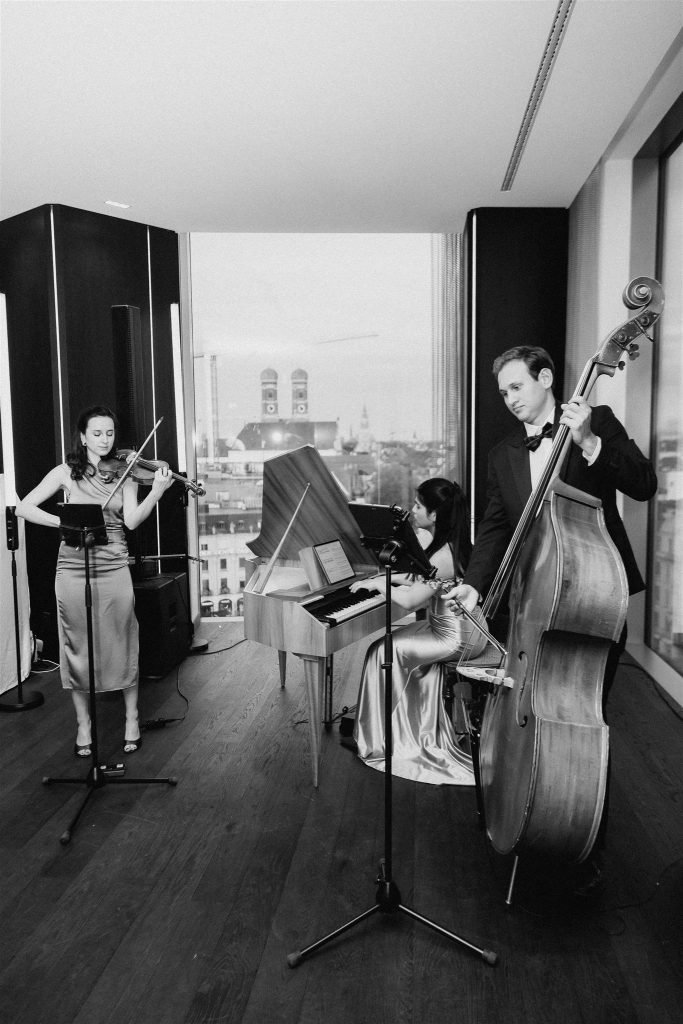 Trio of musicians with violin, piano and double bass in front of the Munich skyline.