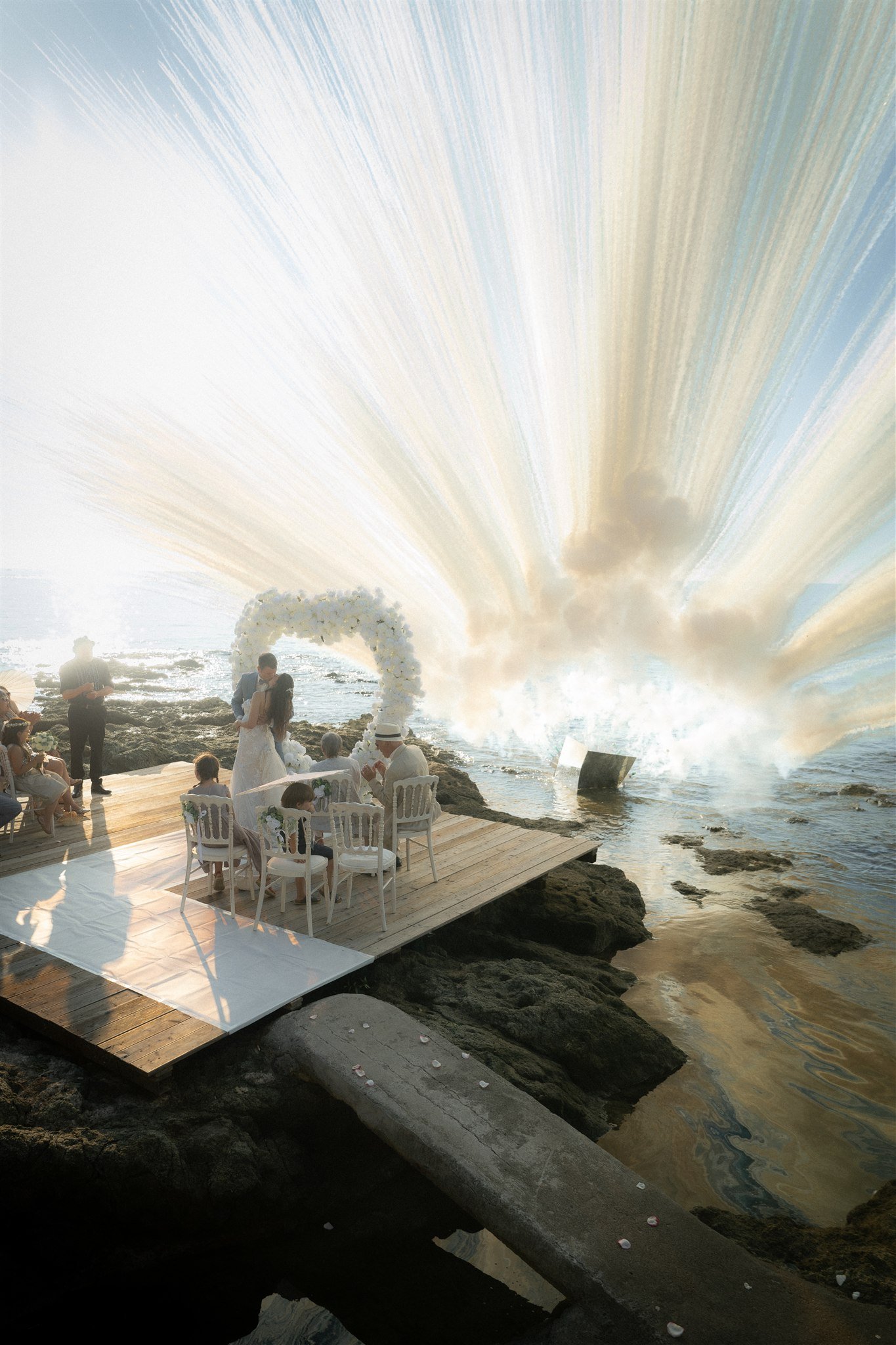First kiss at the seaside ceremony beneath a white floral arch, sun flare over the sea